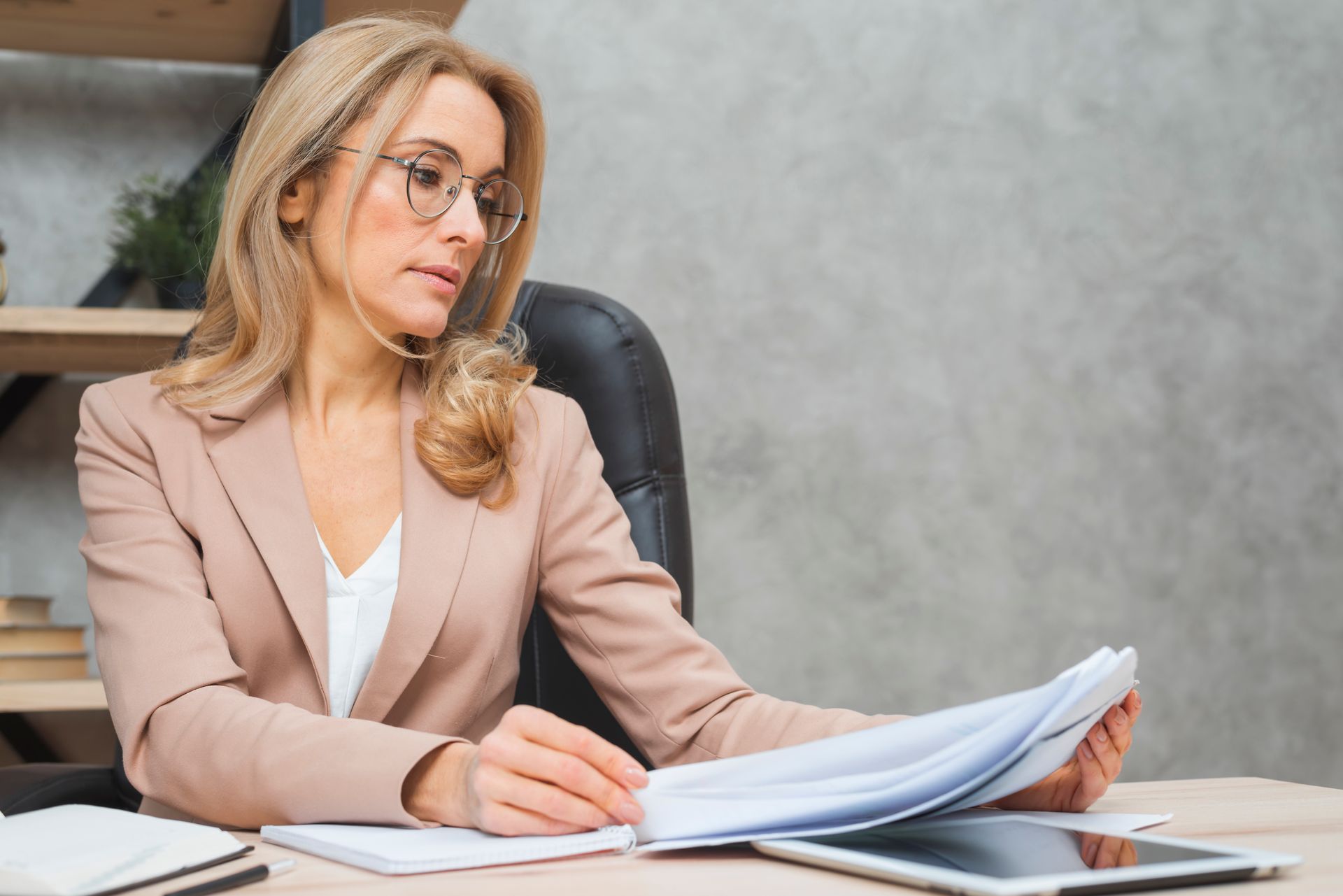 Woman in a beige blazer and glasses reviews documents at a desk in a home office.