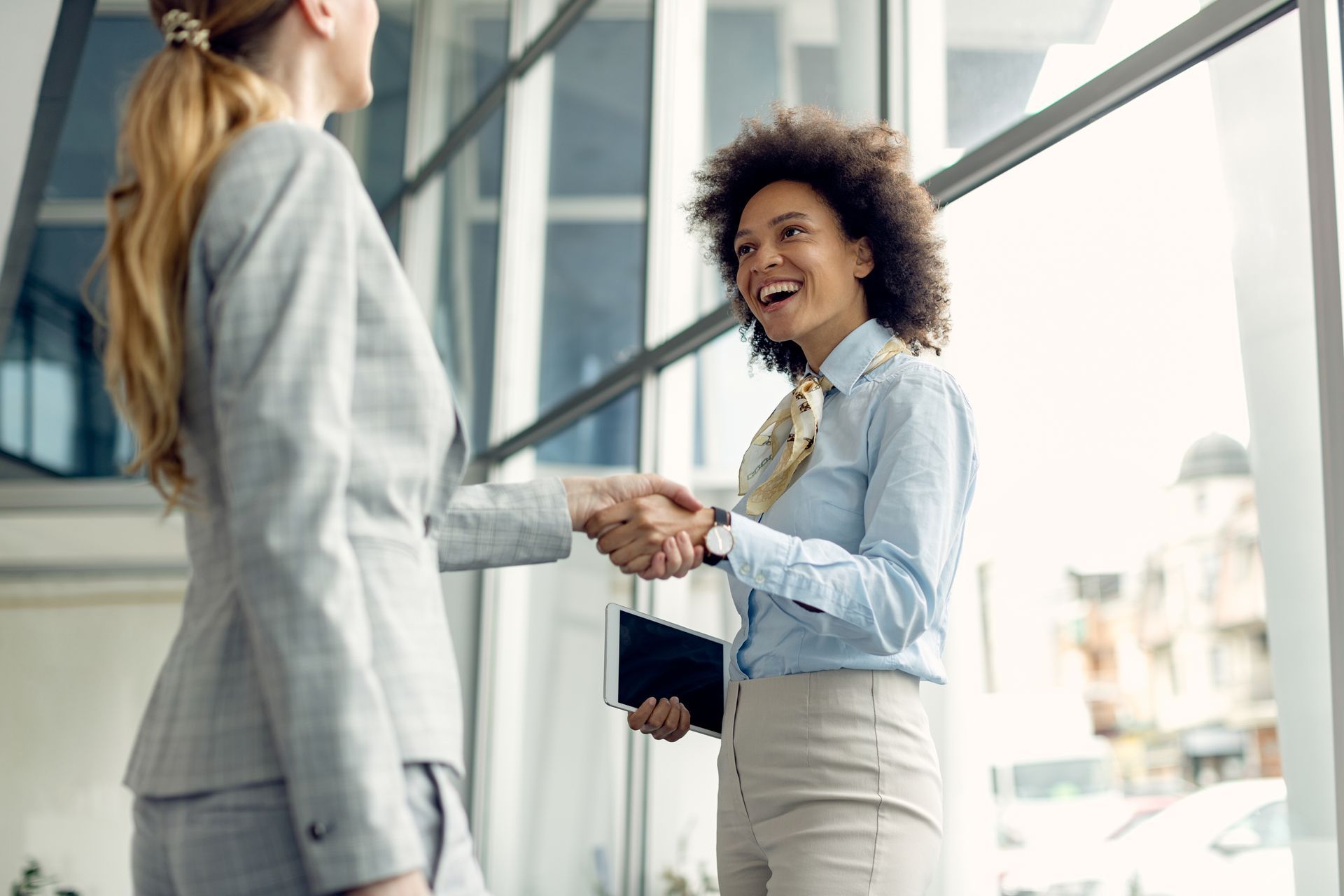 Two businesswomen shaking hands, smiling in a modern office setting.