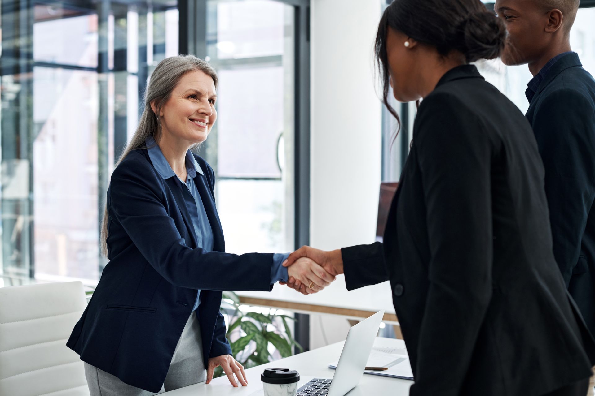 Woman in blazer shakes hands with another woman at a desk in an office setting.