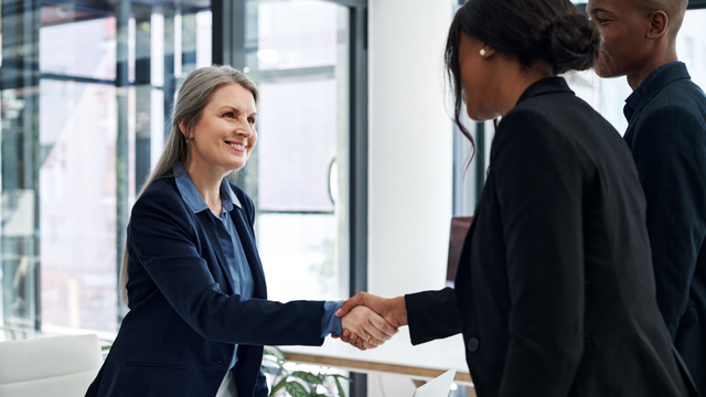 Woman in blue blazer shakes hands with another in a black suit, smiling, in an office.