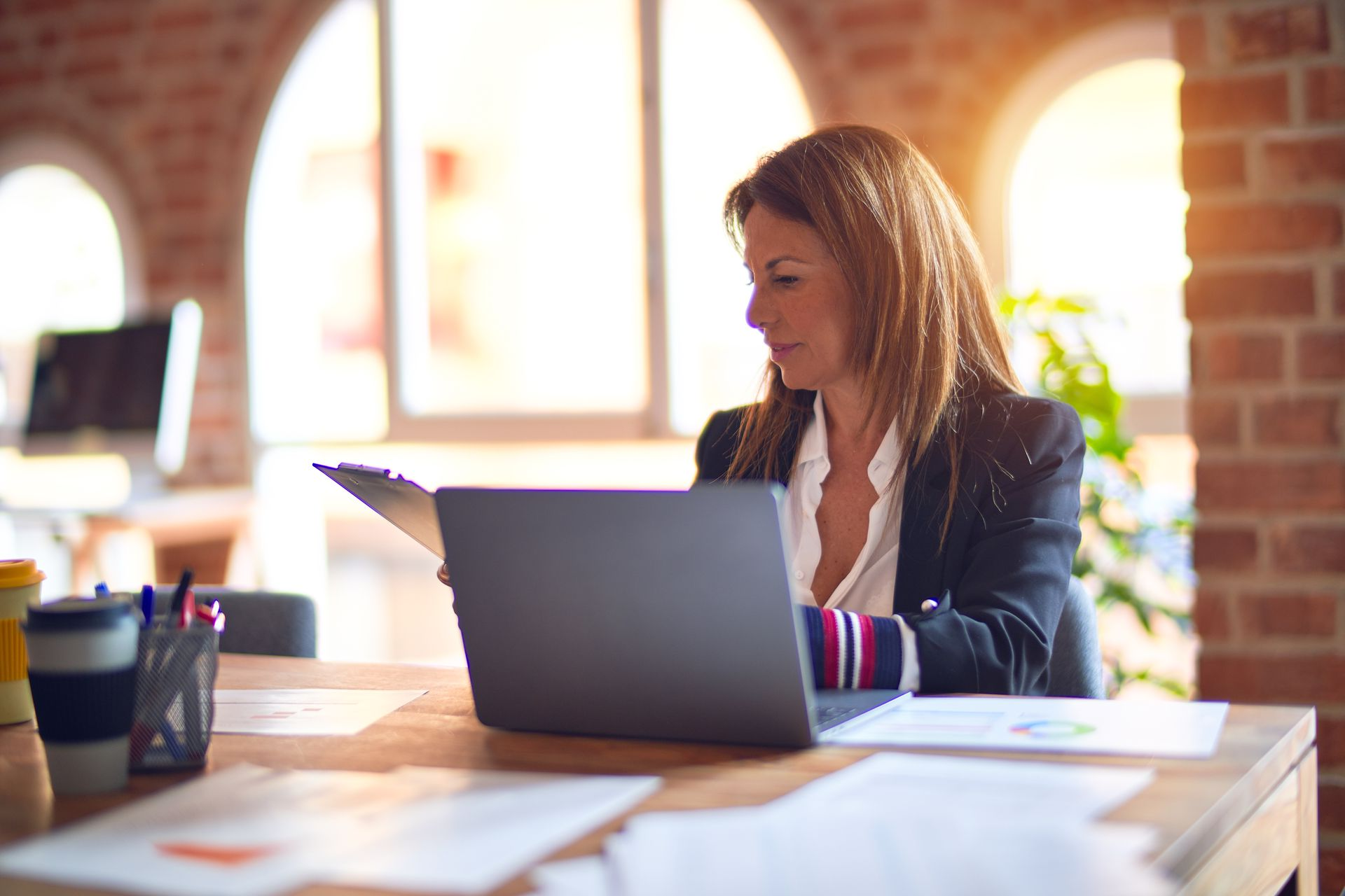 Woman in blazer at desk, looking at tablet, laptop open, brick wall background.