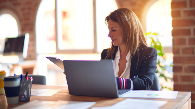 Woman in a blazer working at a desk with a laptop, looking at documents. Sunlight streams in.