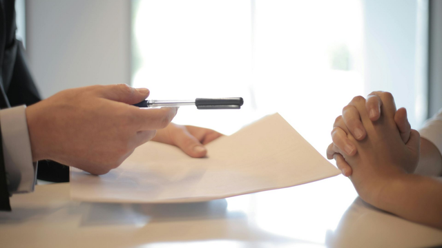 Person in suit handing document and pen to another person; table, bright background.