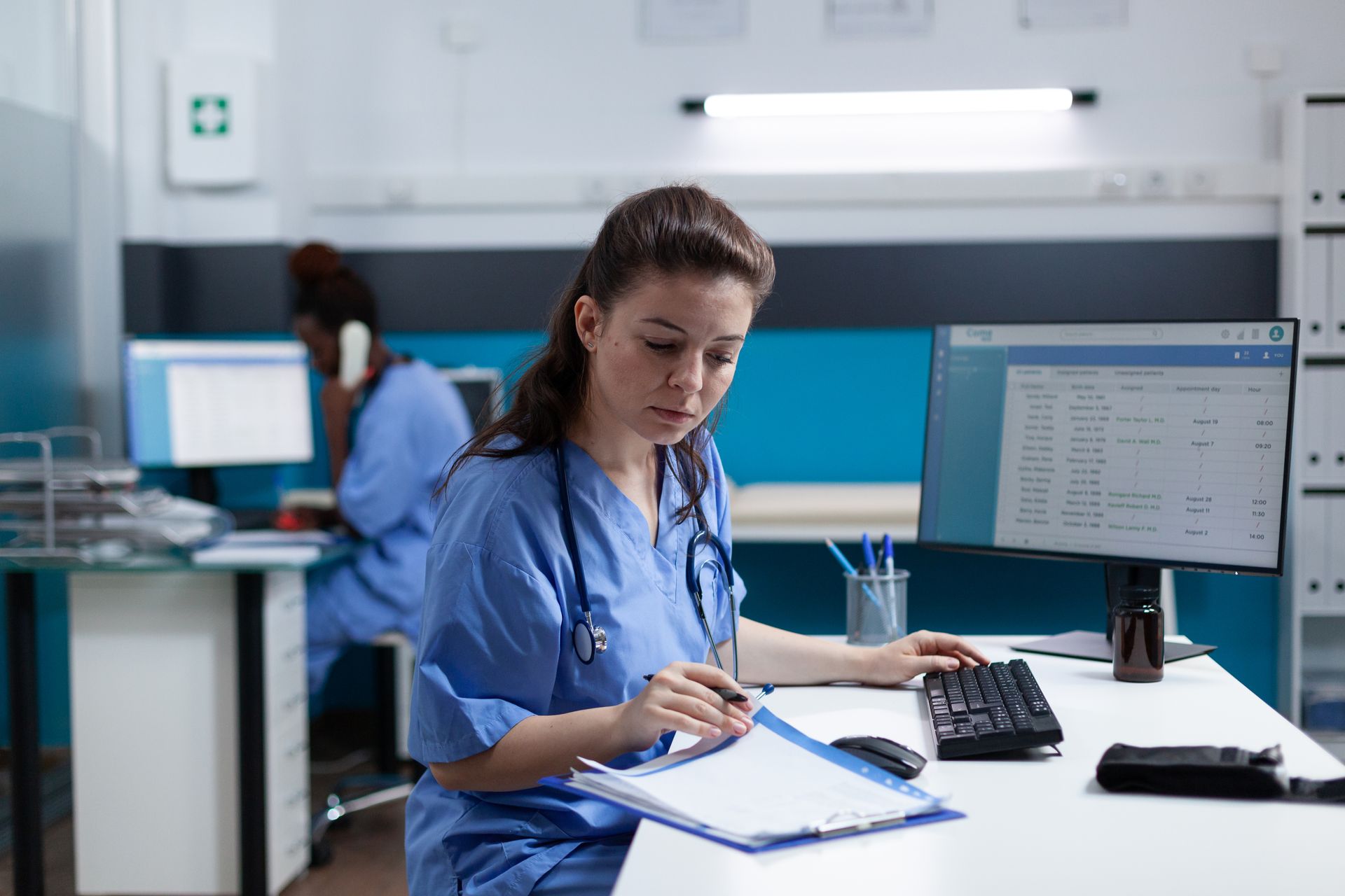 Woman in blue scrubs reviews paperwork at a desk, looking at a computer monitor; another person is in background.