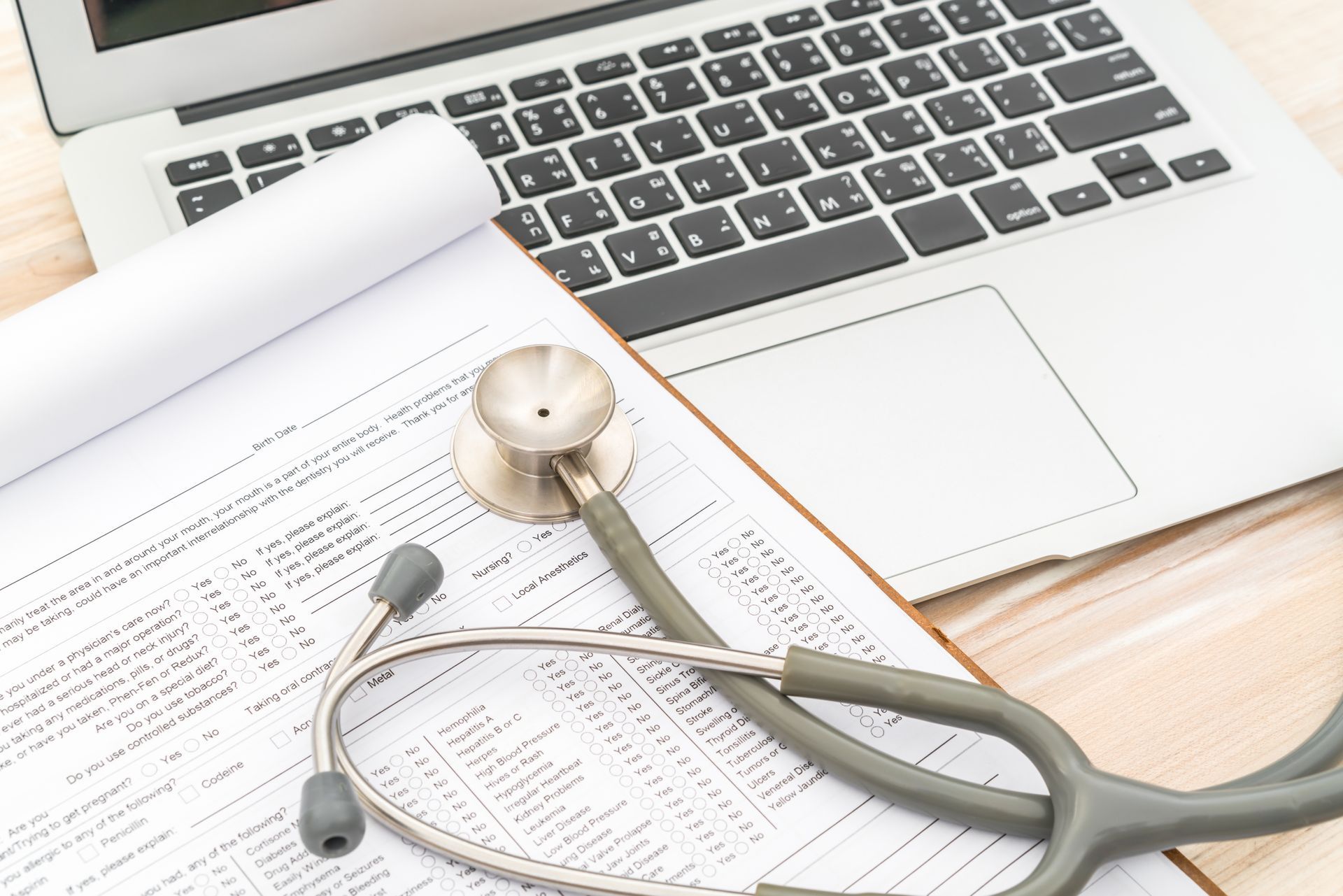 Laptop, clipboard with medical form, and stethoscope on a wooden surface.