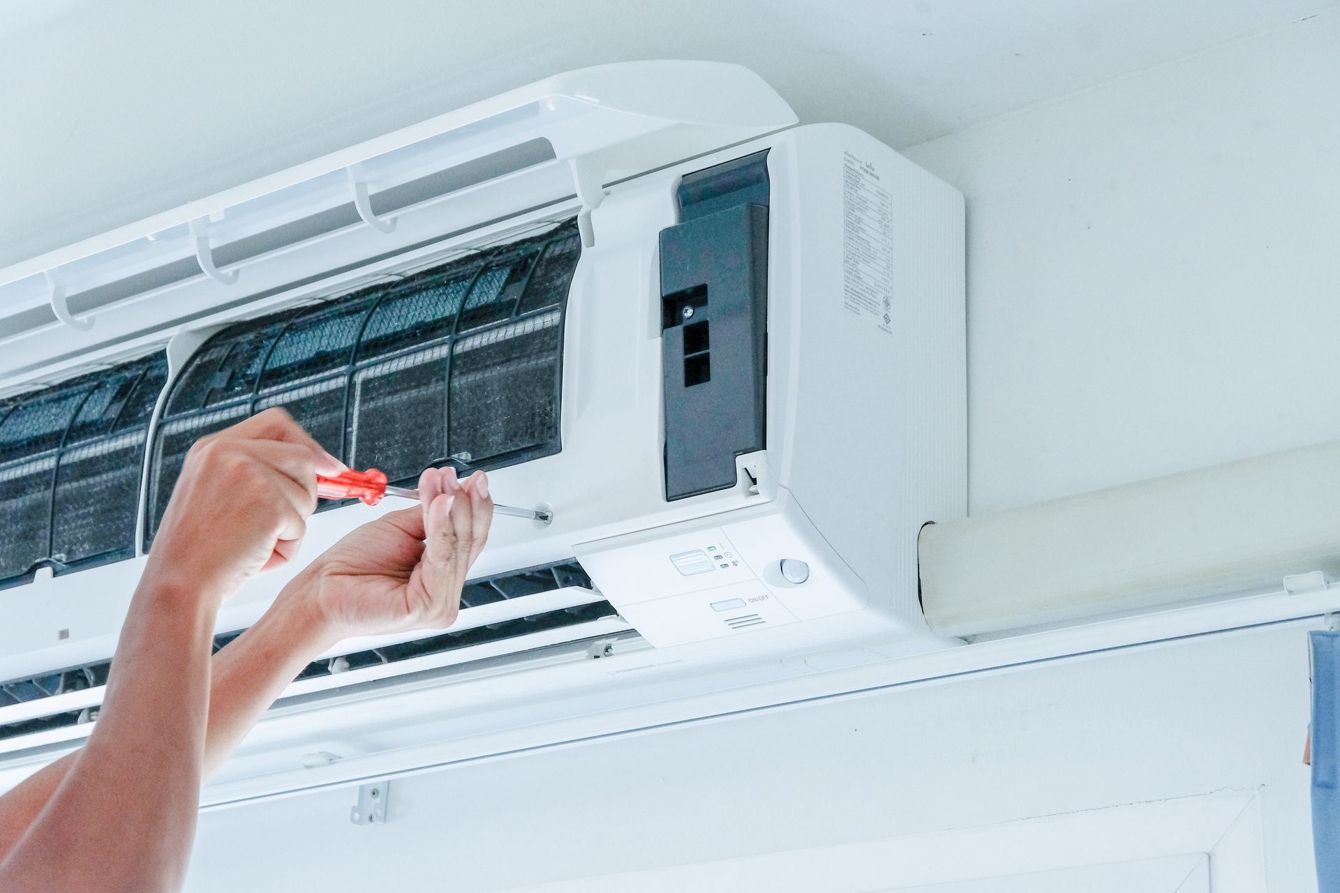 Hands cleaning a white wall-mounted air conditioner with a screwdriver