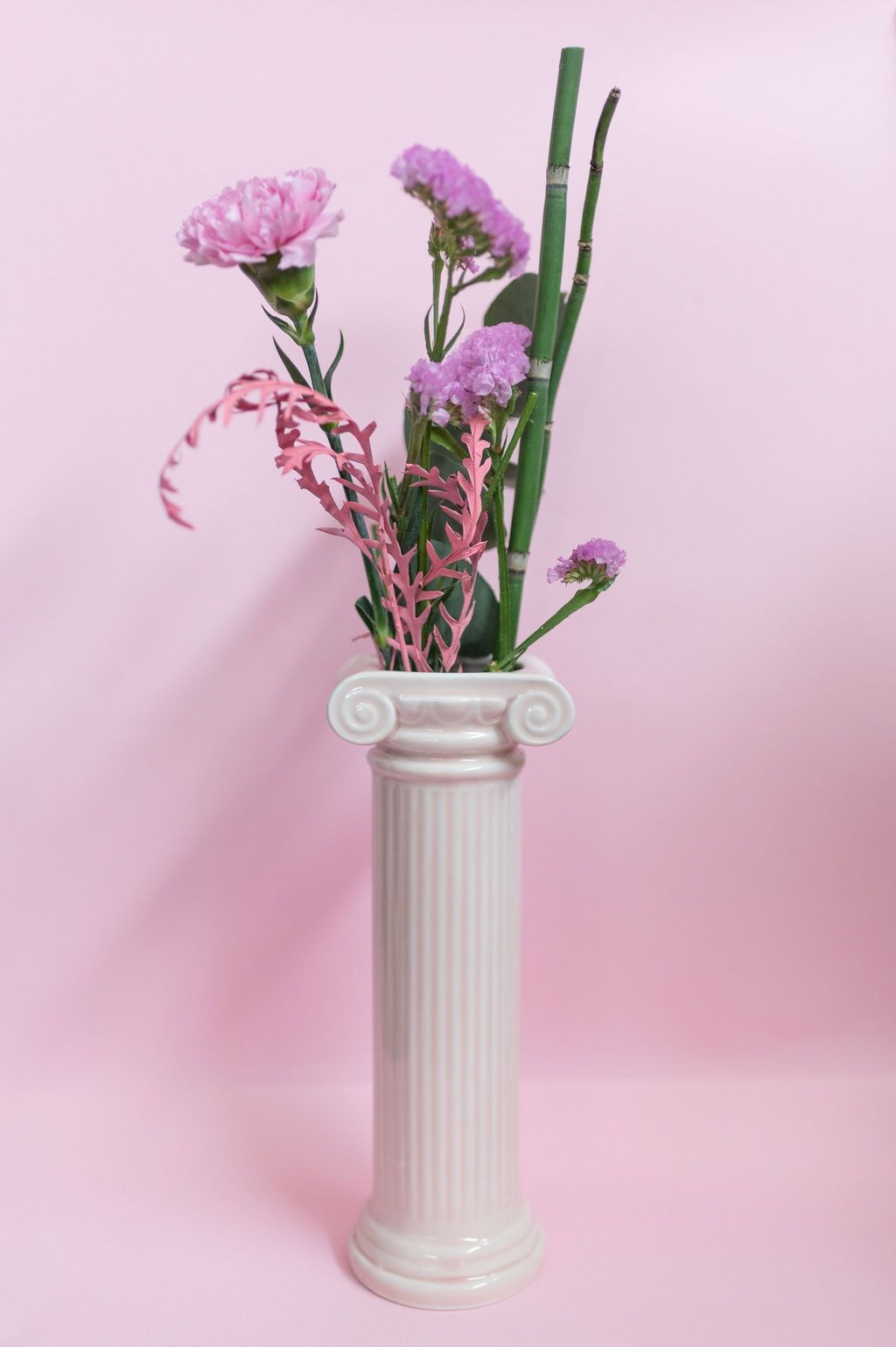 un vaso bianco con fiori viola in cima a una colonna su sfondo rosa .