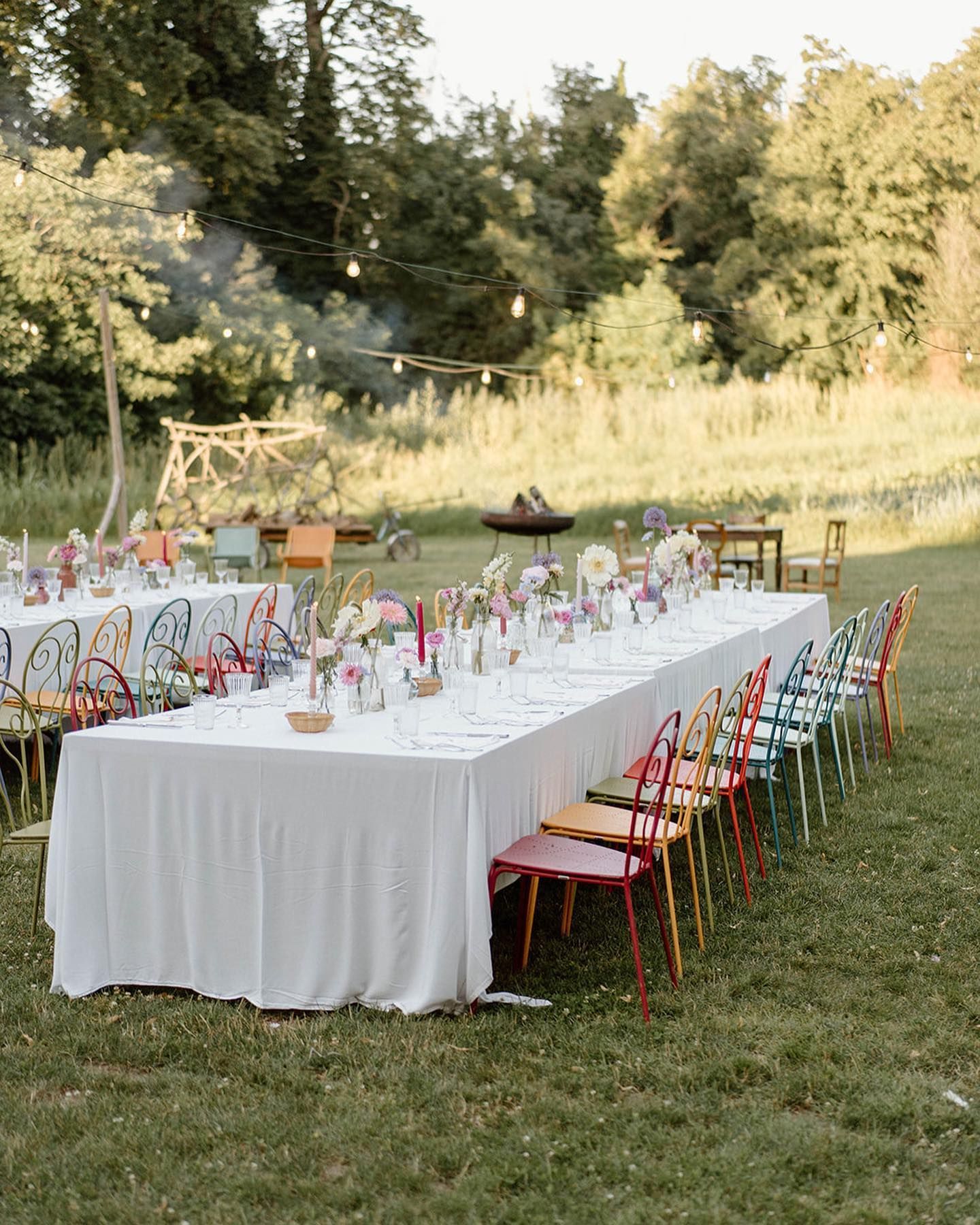un lungo tavolo bianco è seduto nell'erba accanto a una fila di sedie colorate .