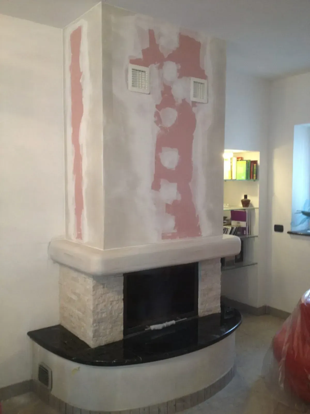 Camino in costruzione, pietra chiara con toppe rosa, focolare nero, pareti bianche.