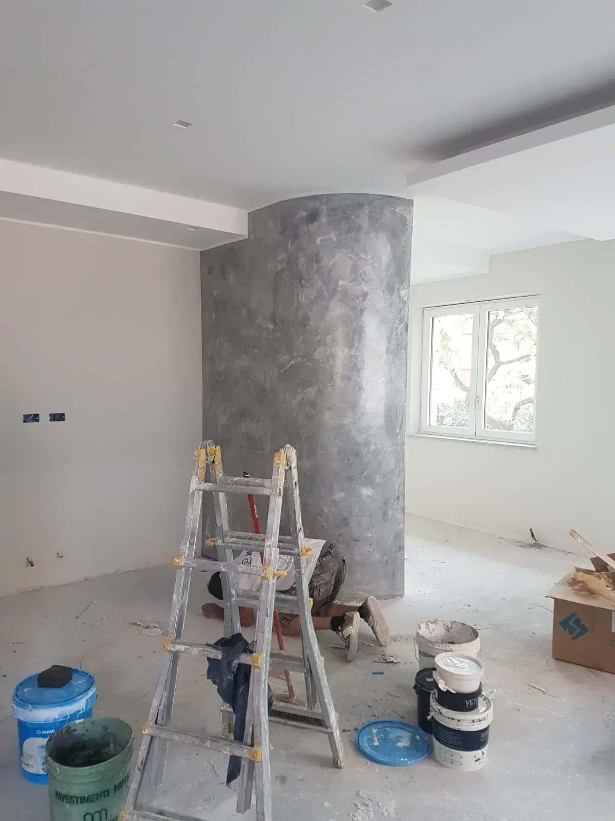 Stanza in fase di ristrutturazione, scala, colonna con finitura grigio screziato, secchi di vernice sul pavimento.
