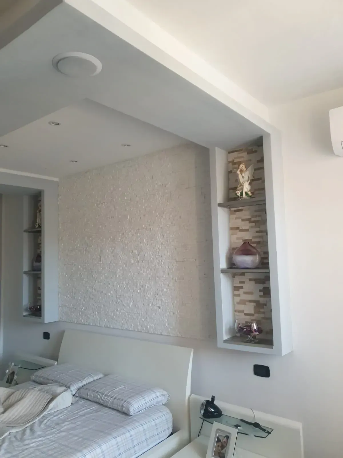 Camera da letto con scaffali integrati; letto bianco, parete strutturata e oggetti decorativi sugli scaffali.