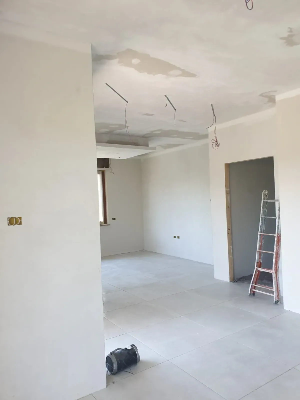Stanza non rifinita con pareti, soffitto e pavimento grigio chiaro. Cavi elettrici pendono dal soffitto, scala sulla porta.