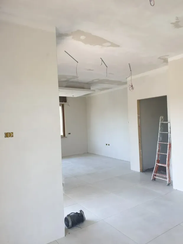 Casa moderna in costruzione con tetto piatto nero e camino in mattoni a vista, circondata da alberi.