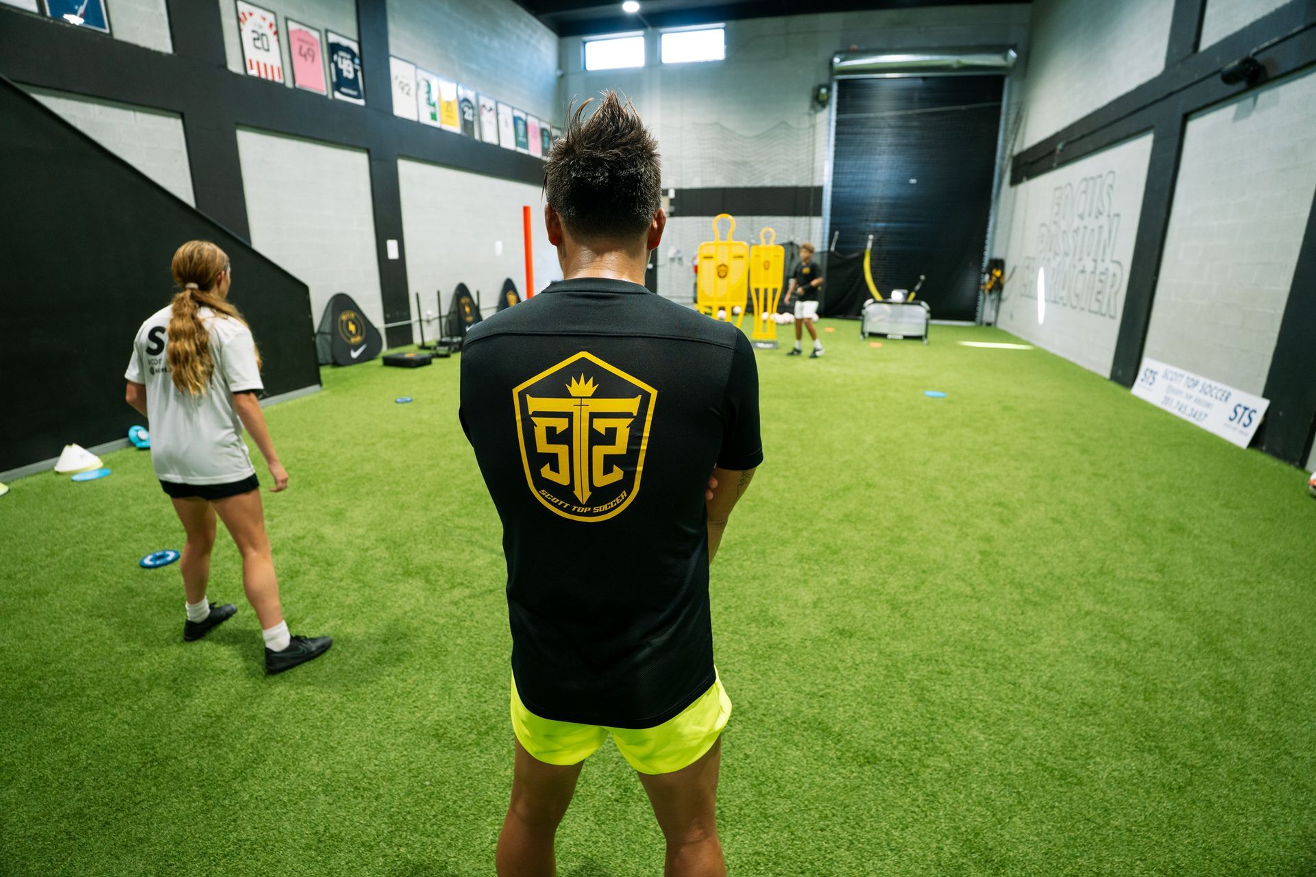 Man in black shirt with logo watching others on artificial turf field.