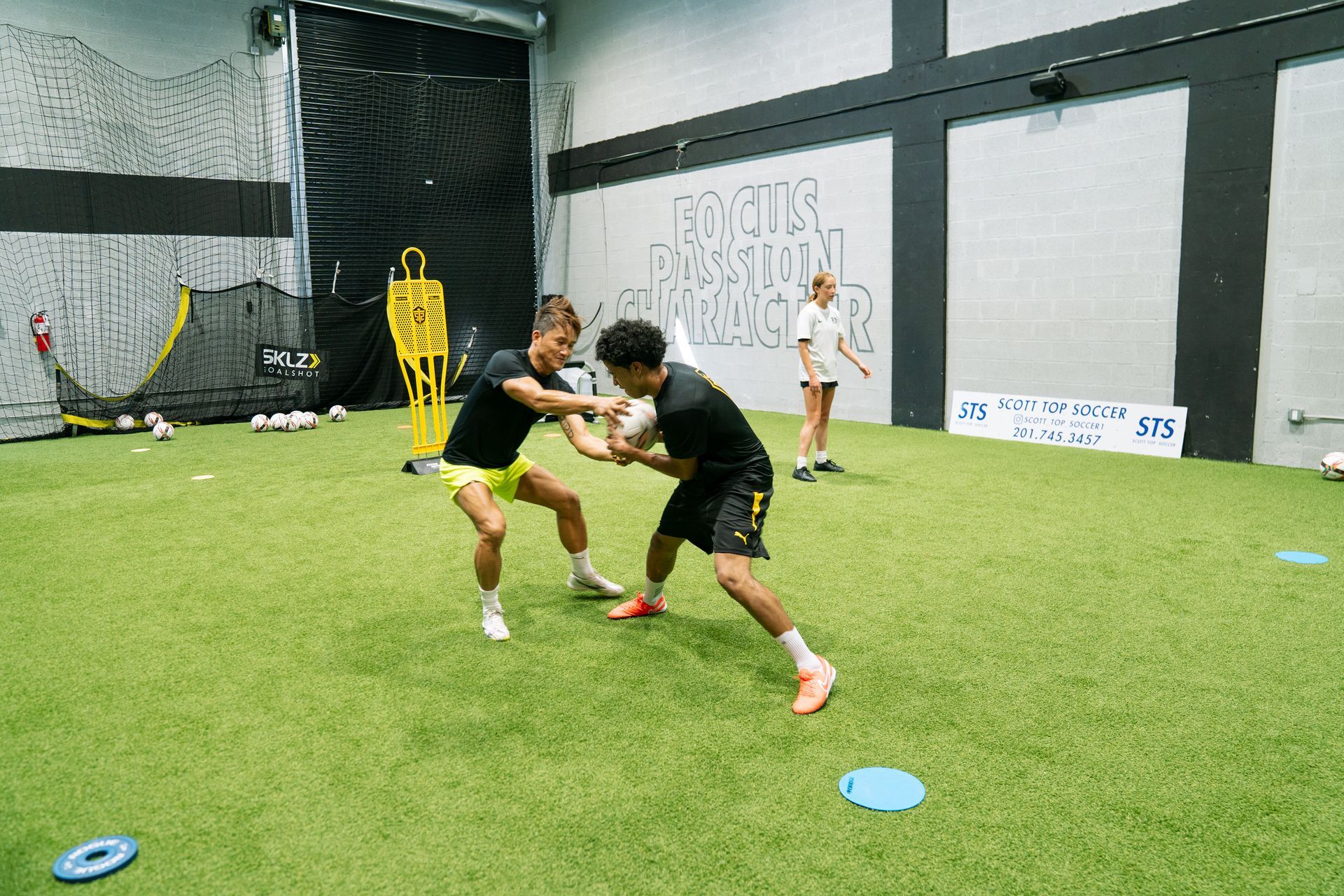 Two athletes in black and yellow practice holding a football on green turf indoors.