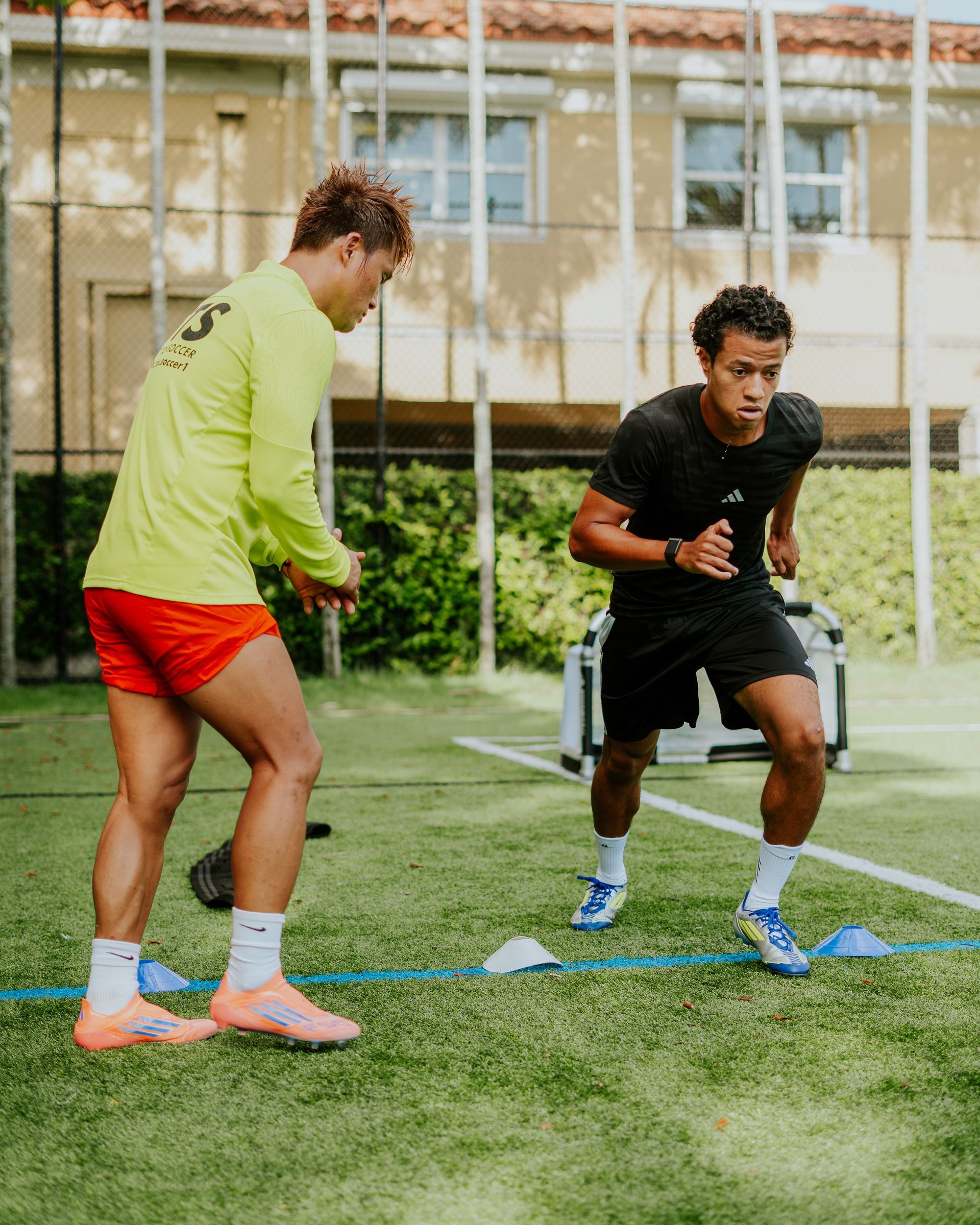 Two men on a green field. One watches the other sprint past cones. Yellow shirt, orange shorts. Black shirt, black shorts.