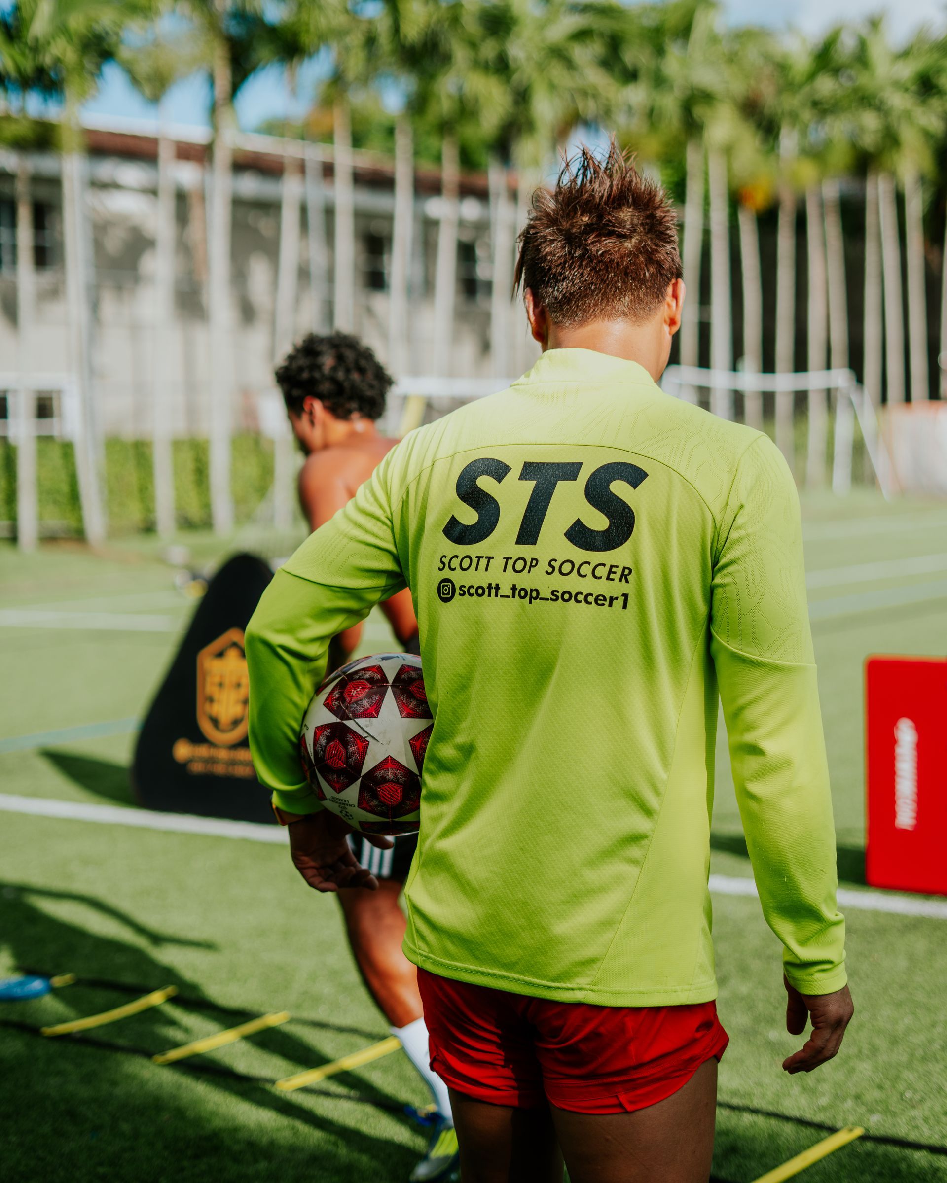 Soccer player in lime green shirt, red shorts holding ball outdoors, training field.