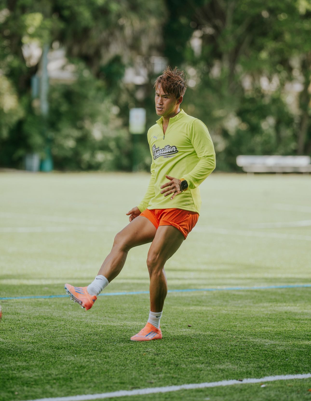 Athlete in orange shorts and yellow top, kicking a ball on a green field.