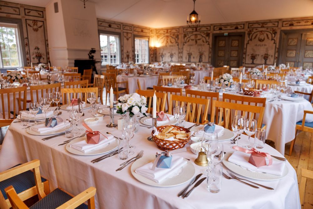 Spisestue dekket til en feiring. Bord dekket med hvitt sengetøy, blomster og gaver. Trestoler og utsmykkede veggmalerier.