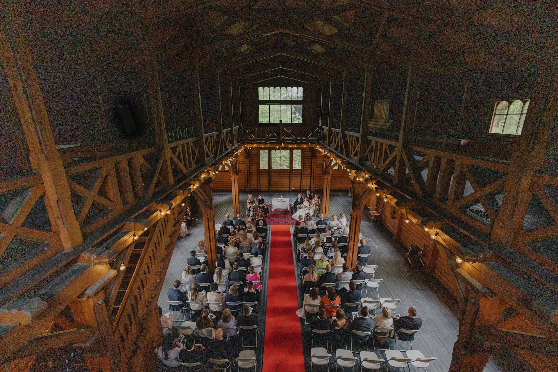 Høyvinkelvisning av en bryllupsseremoni i en rustikk trelåve med gjester sittende på hver side av en rød midtgangsløper.