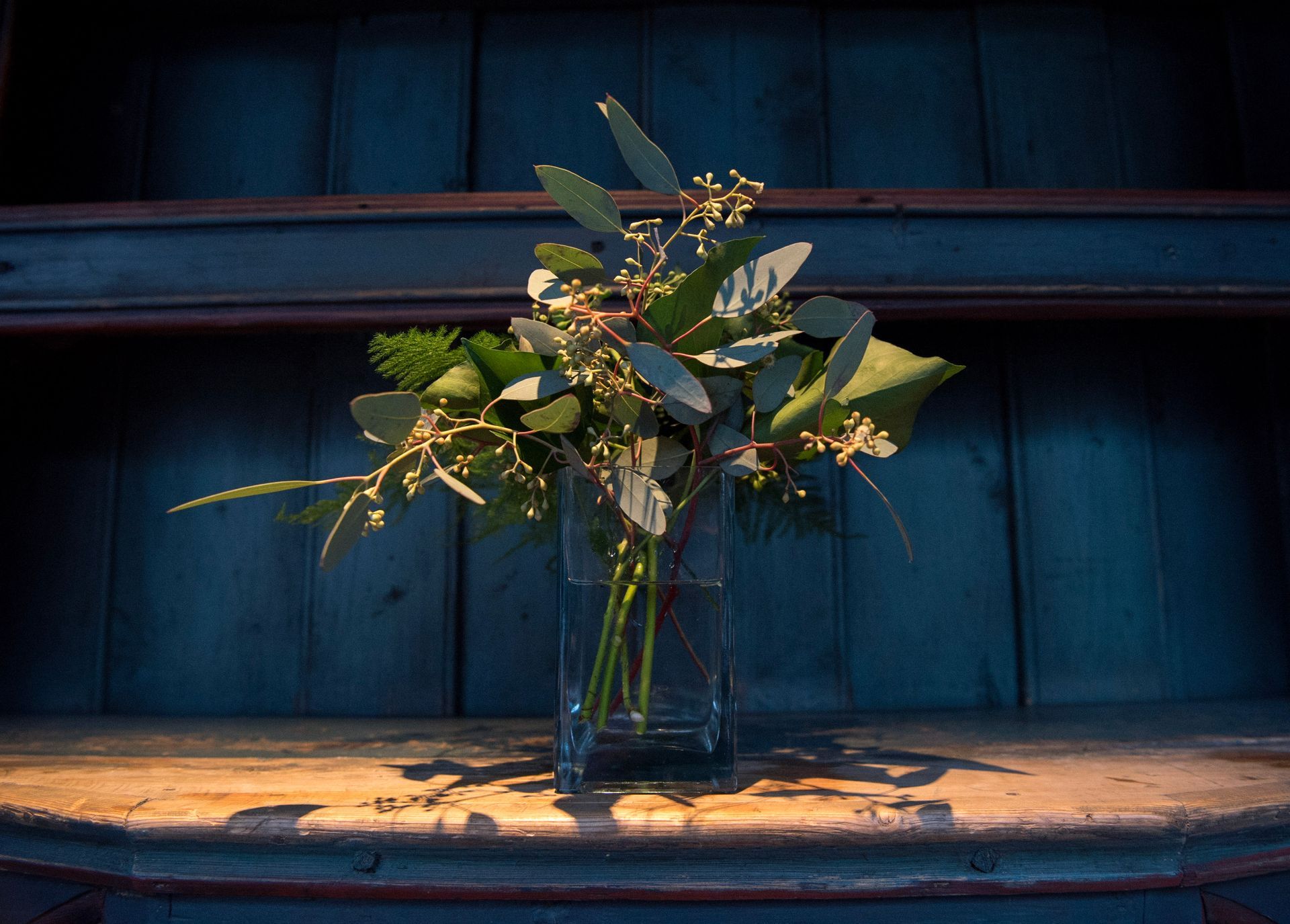 En vase med små blomster på en trehylle, mot en mørkeblå bakgrunn.