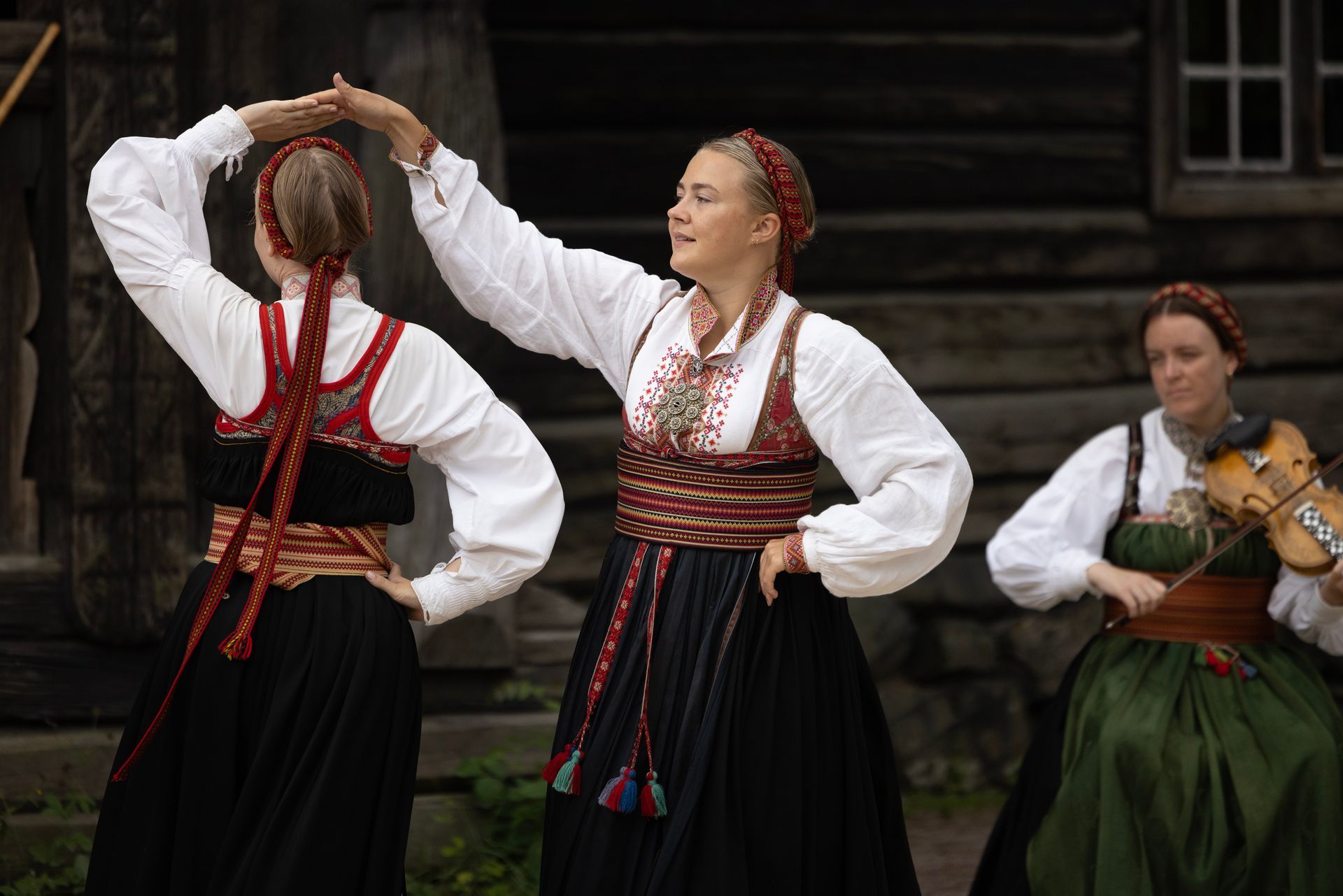 Tre personer i bunad, danser og spiller fiolin utendørs.