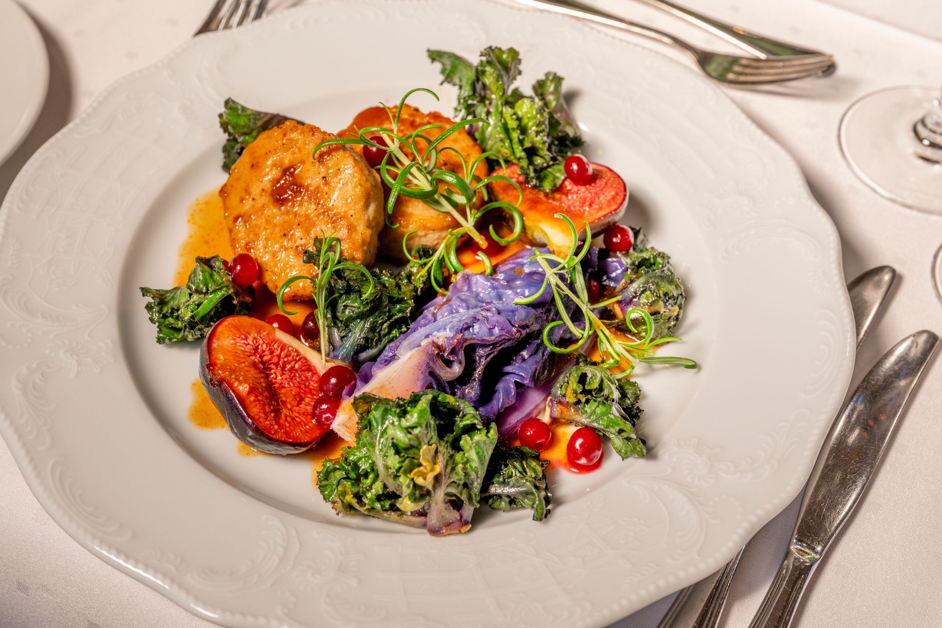 Tallerken med mat med kylling, fiken, grønnkål og lilla kål i en restaurantsetting.