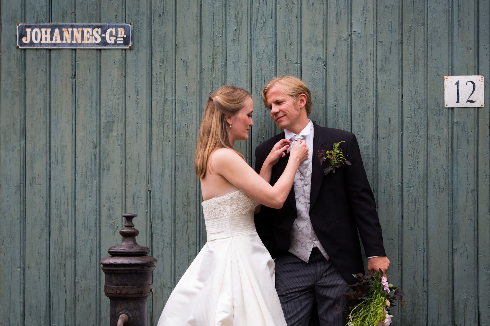 Bruden retter på brudgommens slips, stående ved en grønn tredør med et gateskilt og husnummer 12.