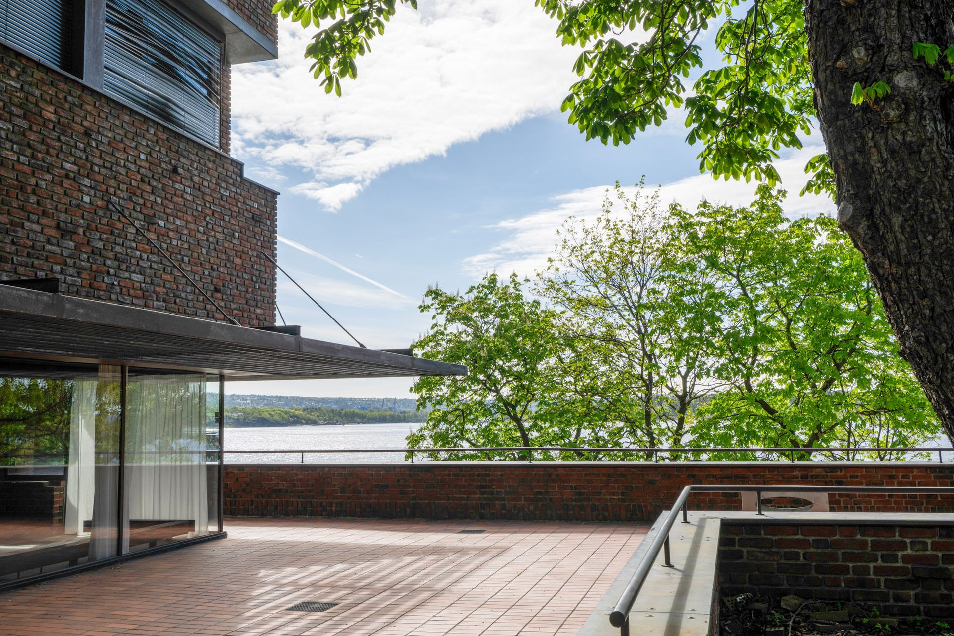 Utvendig utsikt over en murbygning med terrasse, trær og en innsjø under en delvis overskyet himmel.