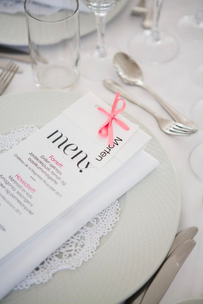 Menykort på en hvit tallerken med sølvtøy, et glass og rosa sløyfe; borddekking.