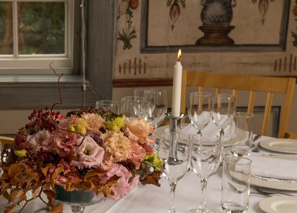 Borddekket til et måltid med et blomsterdekorasjon i midten, tent stearinlys og et vindu i bakgrunnen.