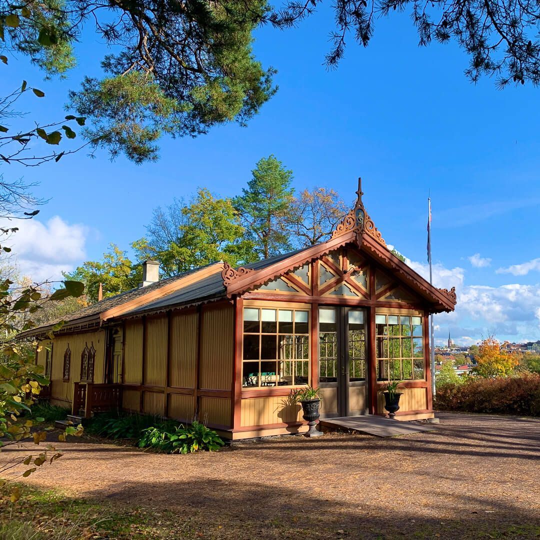 Trebygning med utsmykket tak og store vinduer på en grussti, trær i bakgrunnen, solrik dag.