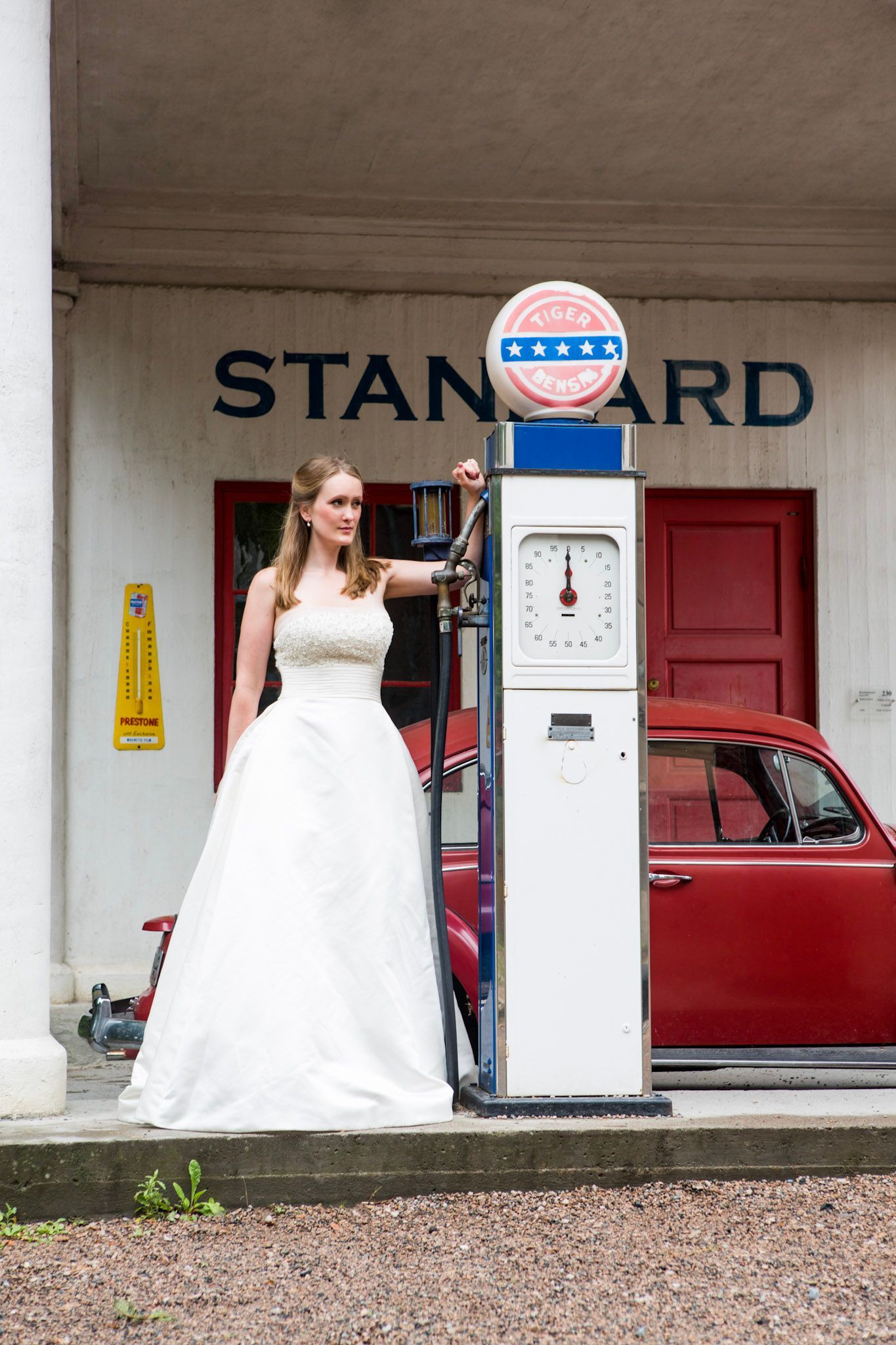 En person i en hvit brudekjole står ved siden av en vintage bensinpumpe på en stasjon med en rød bil i bakgrunnen.