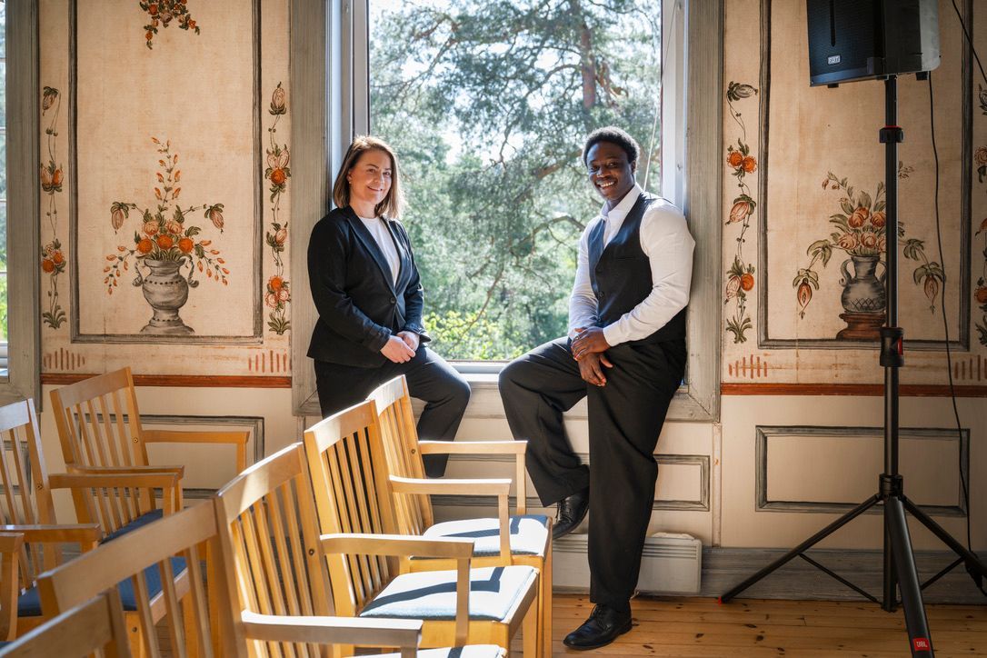 To servitører står ved et vindu i et rom med blomstermalerier på veggene.