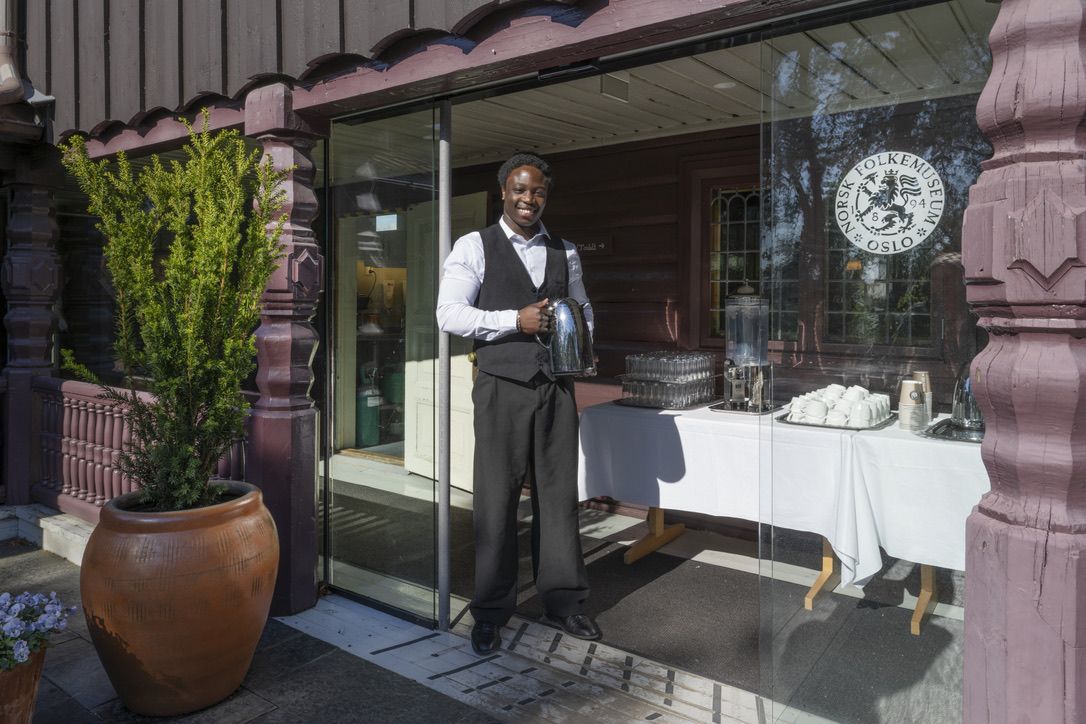 En mann i vest står ved en glassdør i en trebygning og holder en flaske.