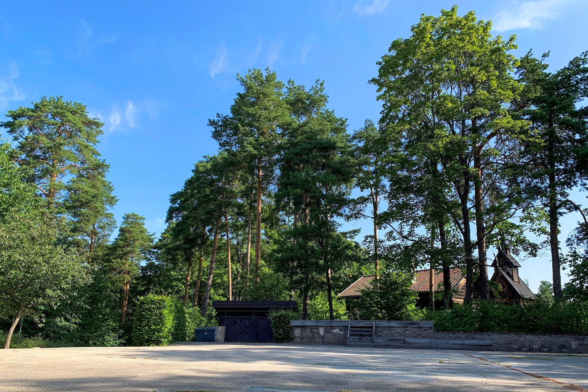 En gruslysning fører til en steinmur, tette grønne trær og en tradisjonell trekonstruksjon mot en knallblå himmel.