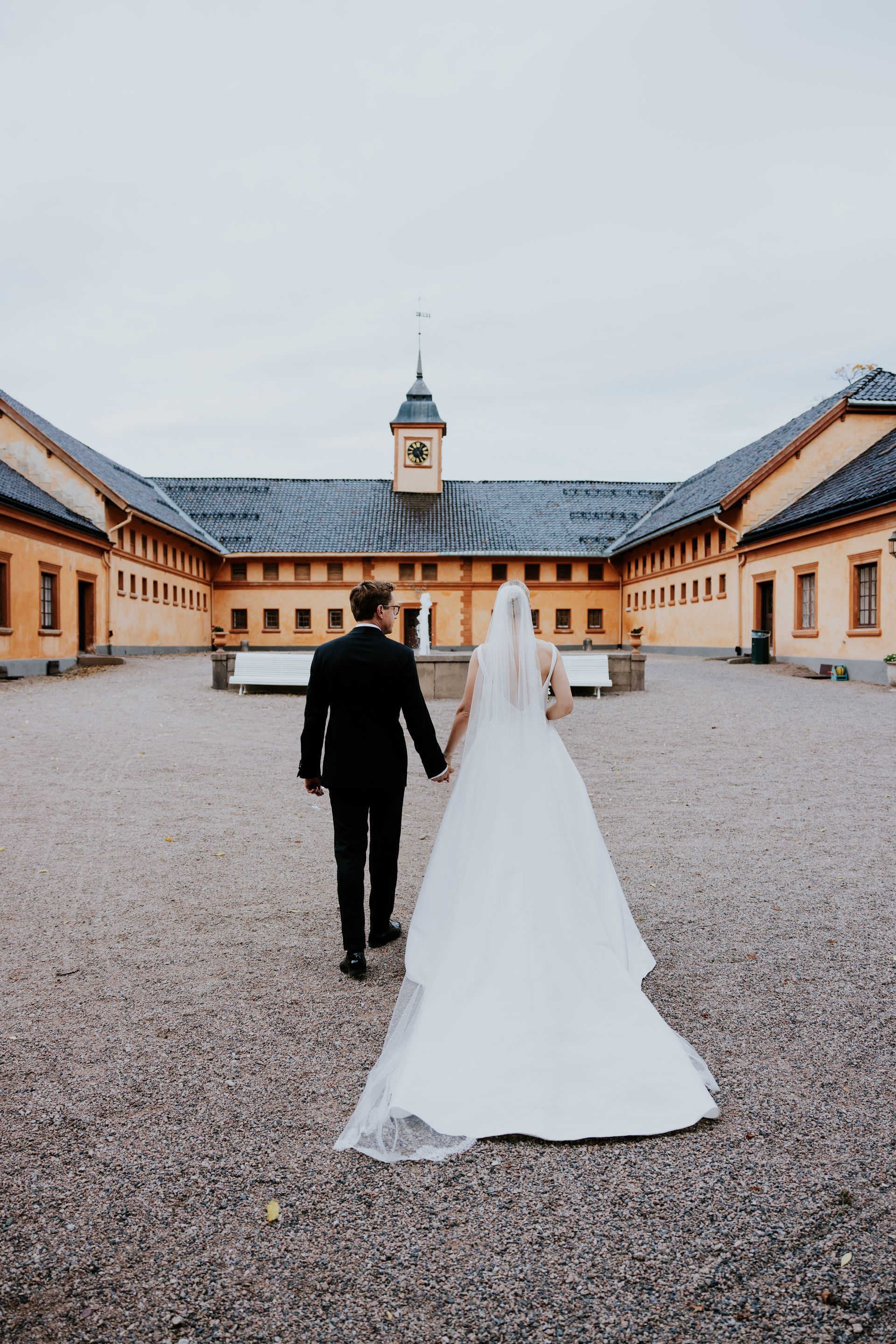 Brud og brudgom går hånd i hånd på gårdsplassen mot bygningen. Brudekjole, slør, dress. Grusunderlag. Overskyet himmel.