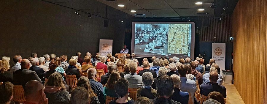 Publikum sitter i en presentasjonssal, ser på en skjerm med bilder og en foredragsholder på et podium.