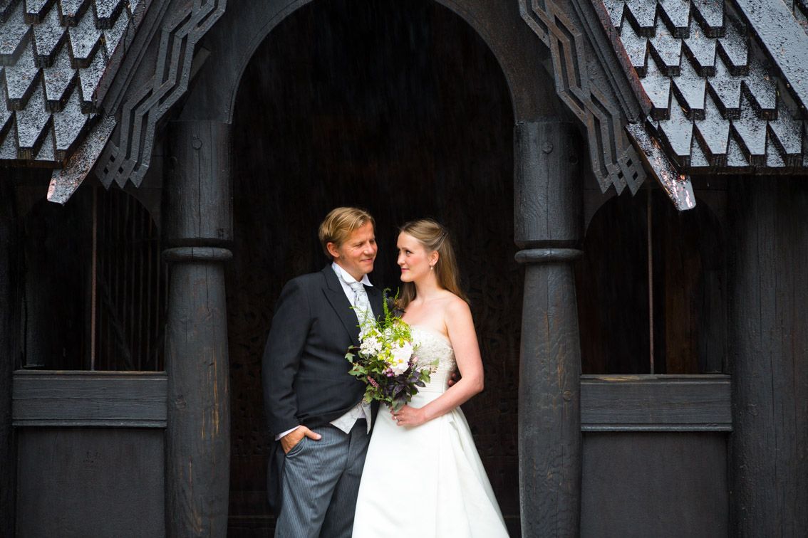 Kvinne med blomsterkrone kysser mannens panne; begge smiler, omgitt av grøntområder.