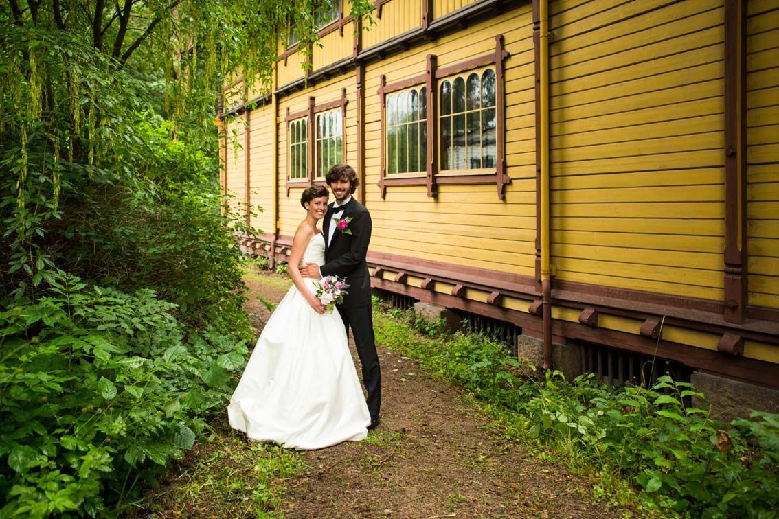 Brud og brudgom omfavner hverandre ved en gul bygning med utsmykkede vinduer, omgitt av grønt løvverk.