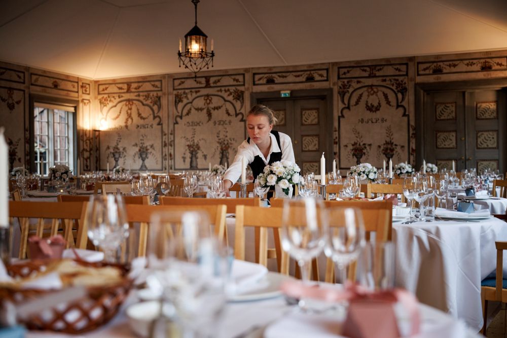 En servitør arrangerer blomster på et bord dekket til et formelt arrangement i et rom med utsmykket veggdekor.