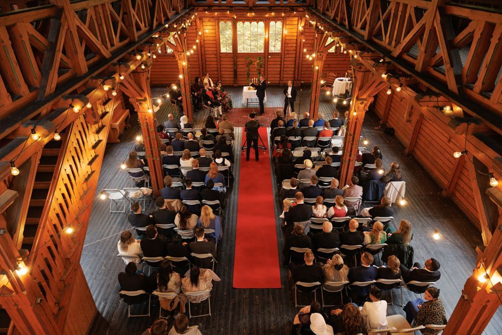 Bryllupsseremoni i bygning med trebjelker. Rød løper i midtgangen, rekker av gjester, lyslenker.