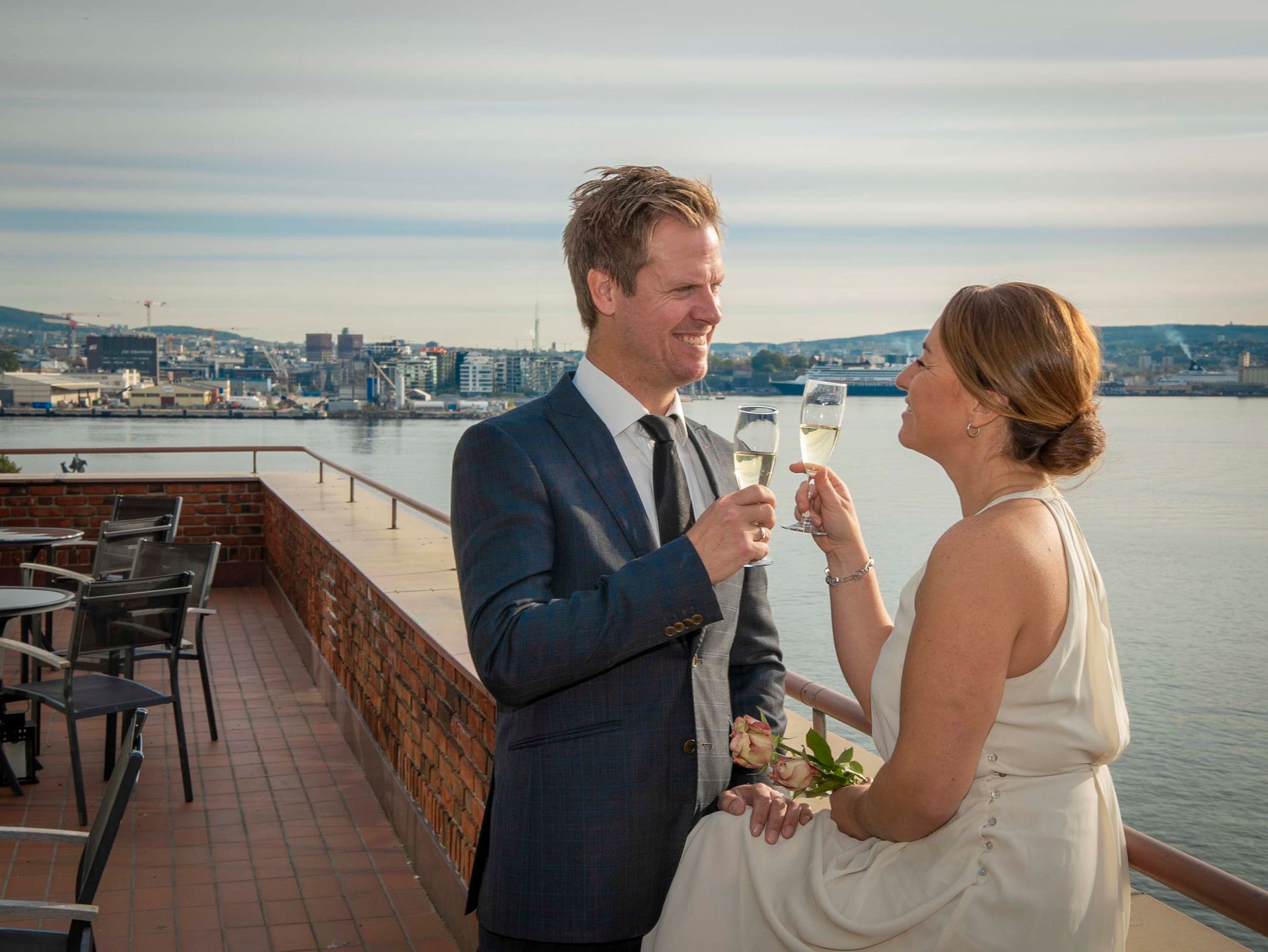 Par klirrer med champagneglass på en balkong med utsikt over en by og vann.