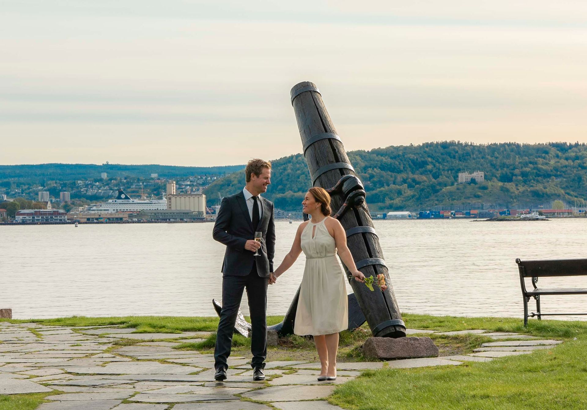 Paret holder hender, smiler og går nær en kanon med utsikt over et vann og en fjern by.