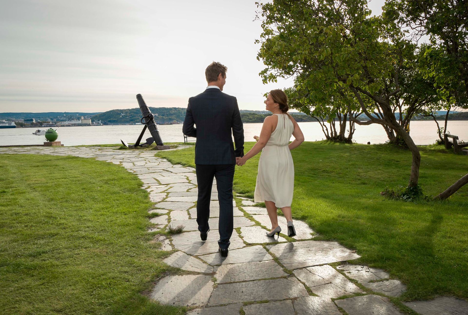 Par går hånd i hånd på en steinsti mot vannet, solrik dag.