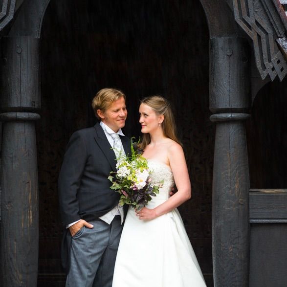 Brudepar, mann i dress og kvinne i hvit kjole, smiler til hverandre, står i en mørk døråpning med tresøyler.