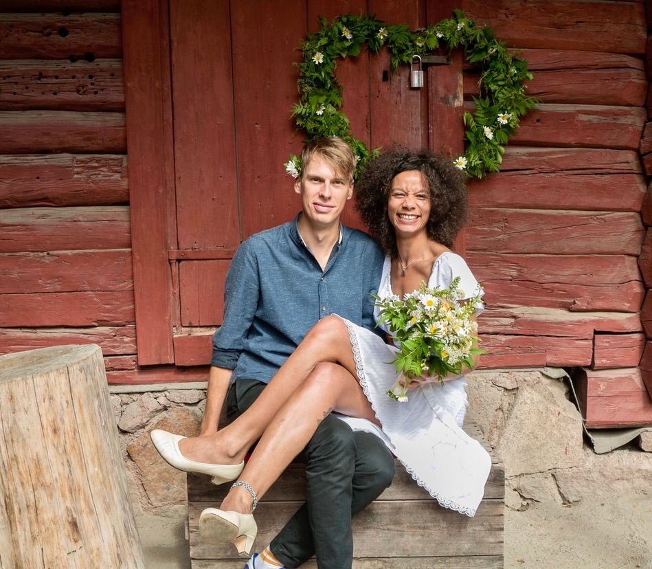 Par sittende; kvinne i hvit kjole, mann i blått, nær en rød trebygning med en hjerteformet krans.