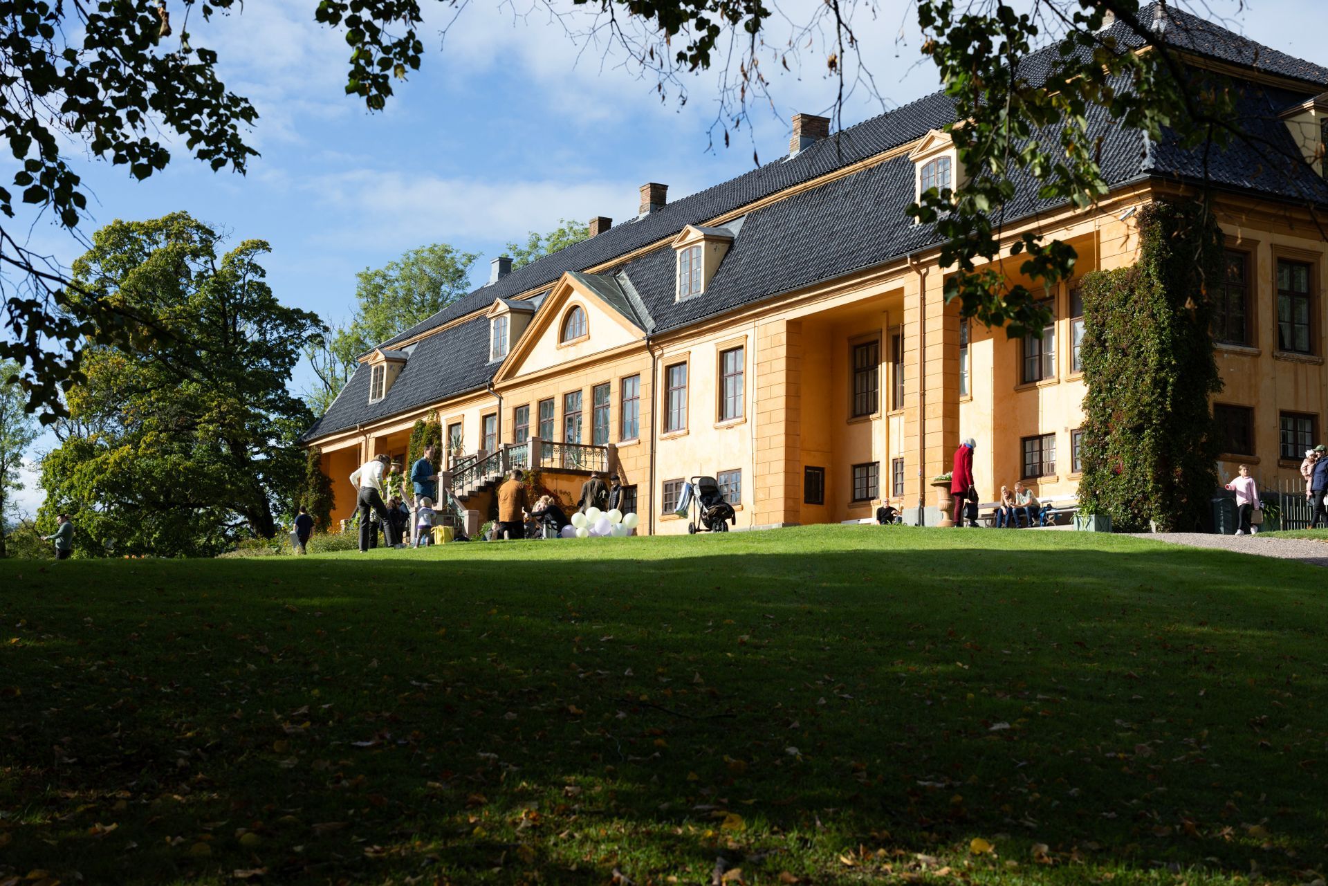 Gul herskapsbolig med mørkt tak og plen. Folk er samlet på en balkong og plenen på en solrik dag.