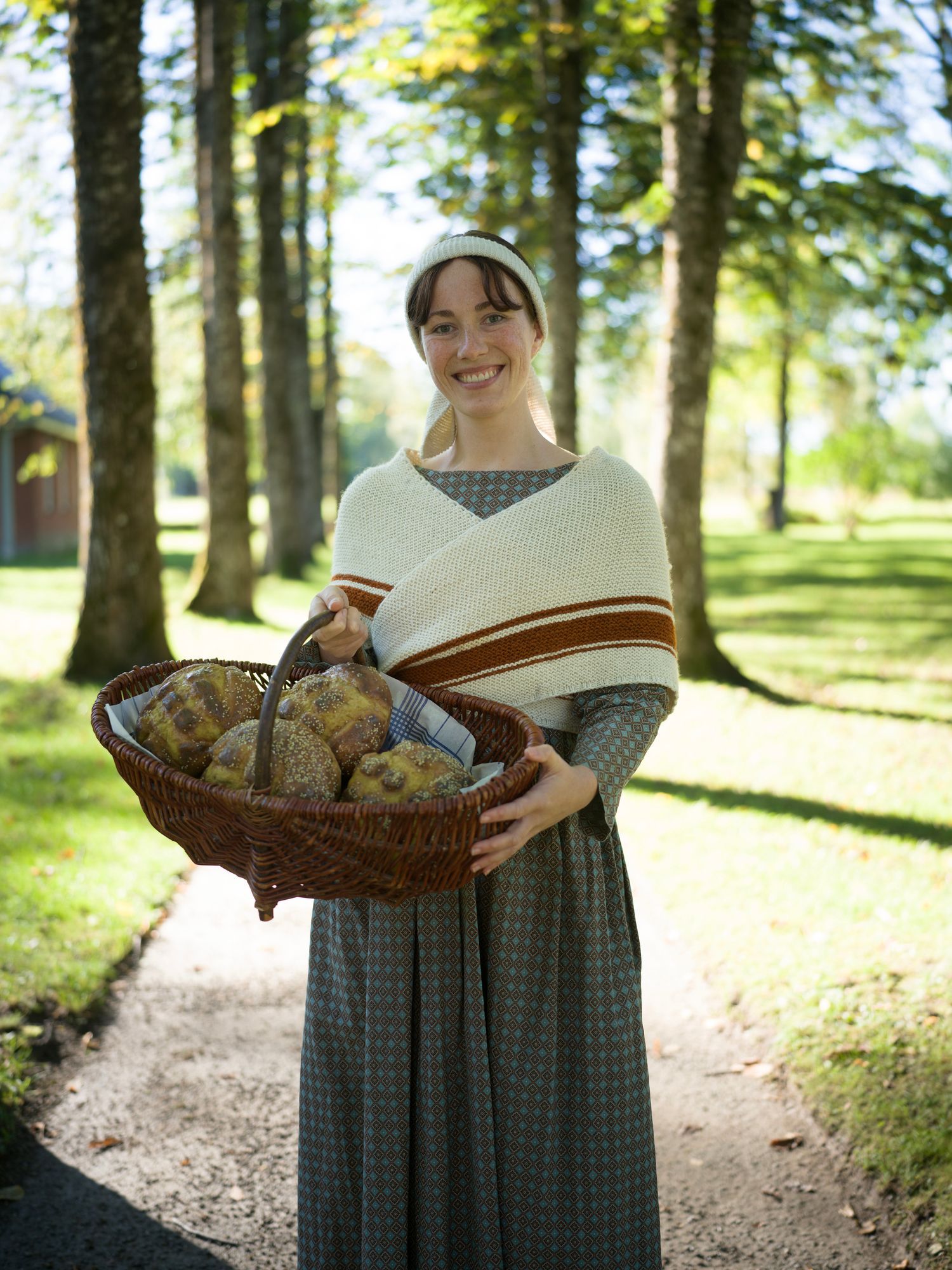 Kvinne i periodekjole bærer brødkurv på en sti mellom trær.