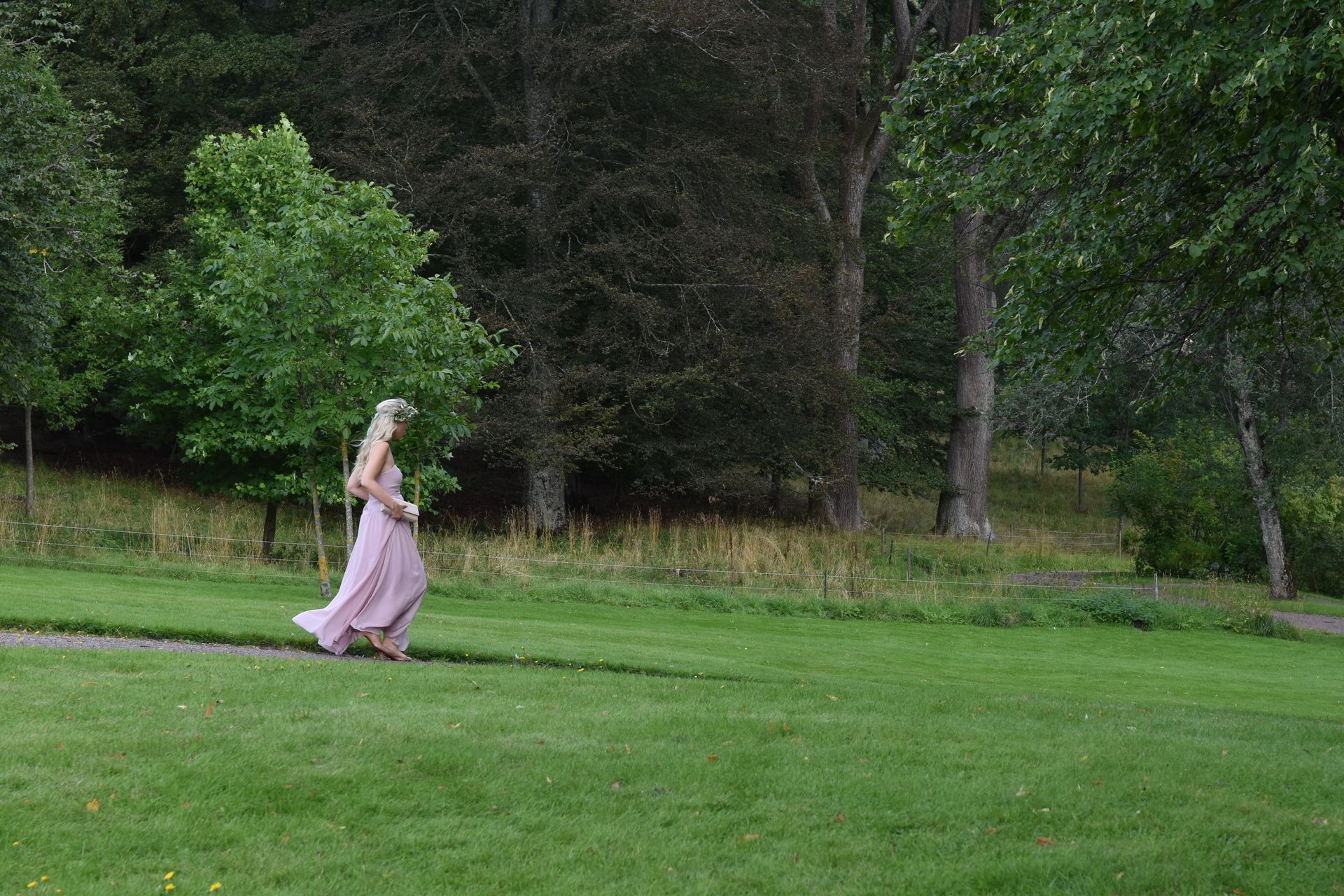 Kvinne i rosa kjole går på en gresskledd sti i en park, med trær i bakgrunnen.
